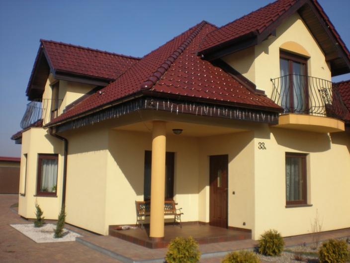 Żółty dom jednorodzinny z brązowym dachem, balkonem z kutą balustradą i numerem 33 na ścianie.