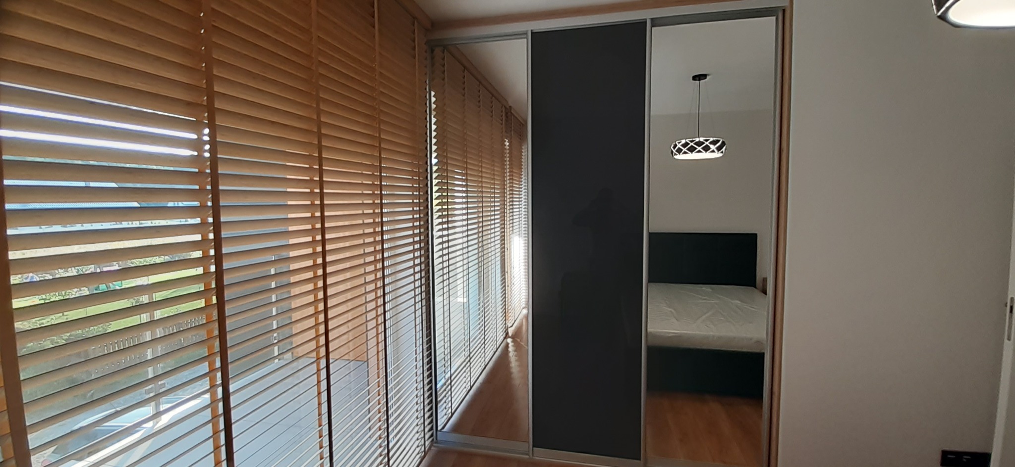 Wnętrze sypialni z przesuwnymi drzwiami szafy z lustrem i czarnym panelem, widok przez drewniane żaluzje okienne.