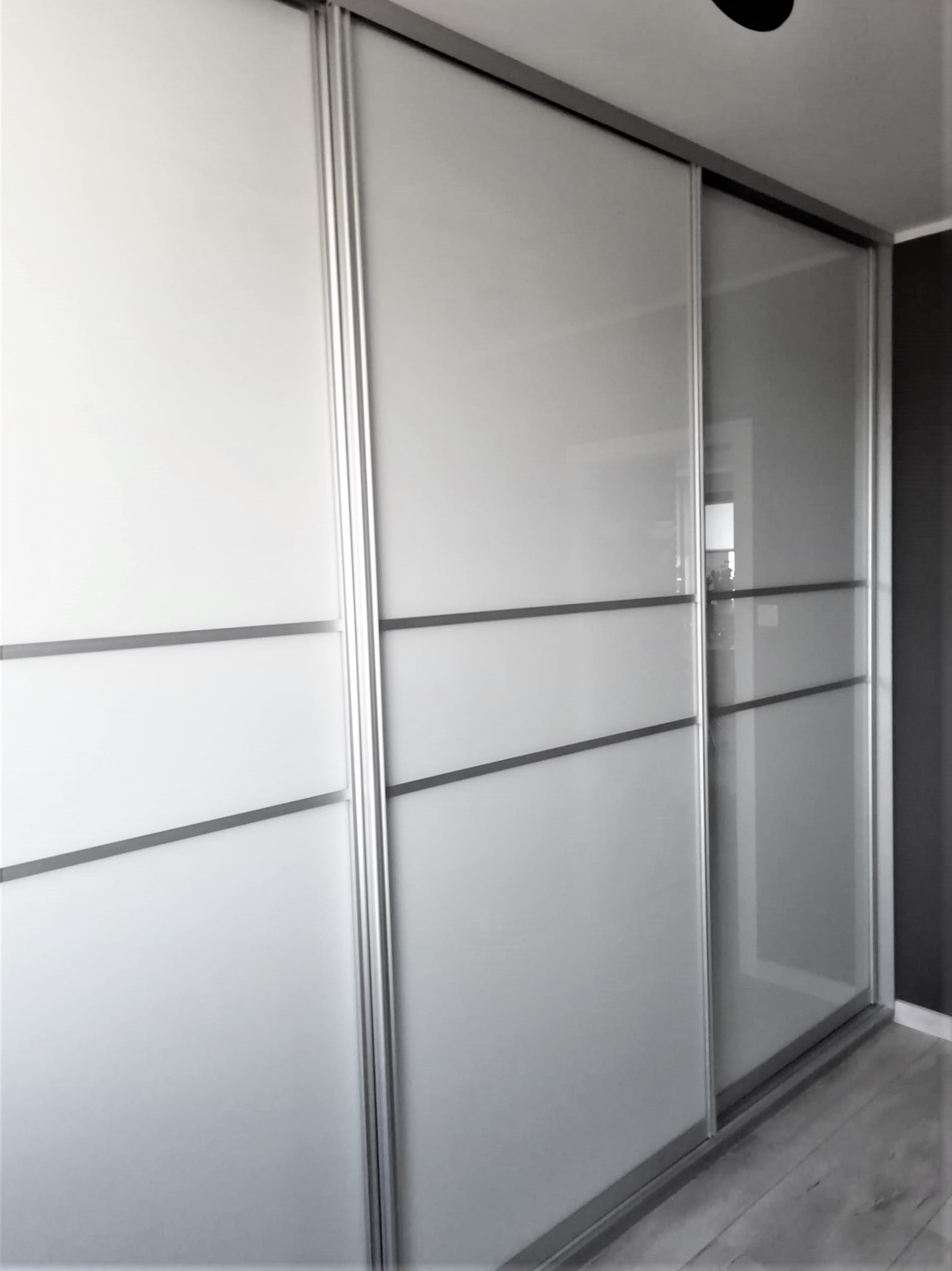 Trzydrzwiowa, biała garderoba z przesuwnymi drzwiami i poziomymi, szarymi akcentami, widoczna na tle szarej ściany i drewnianej podłogi.