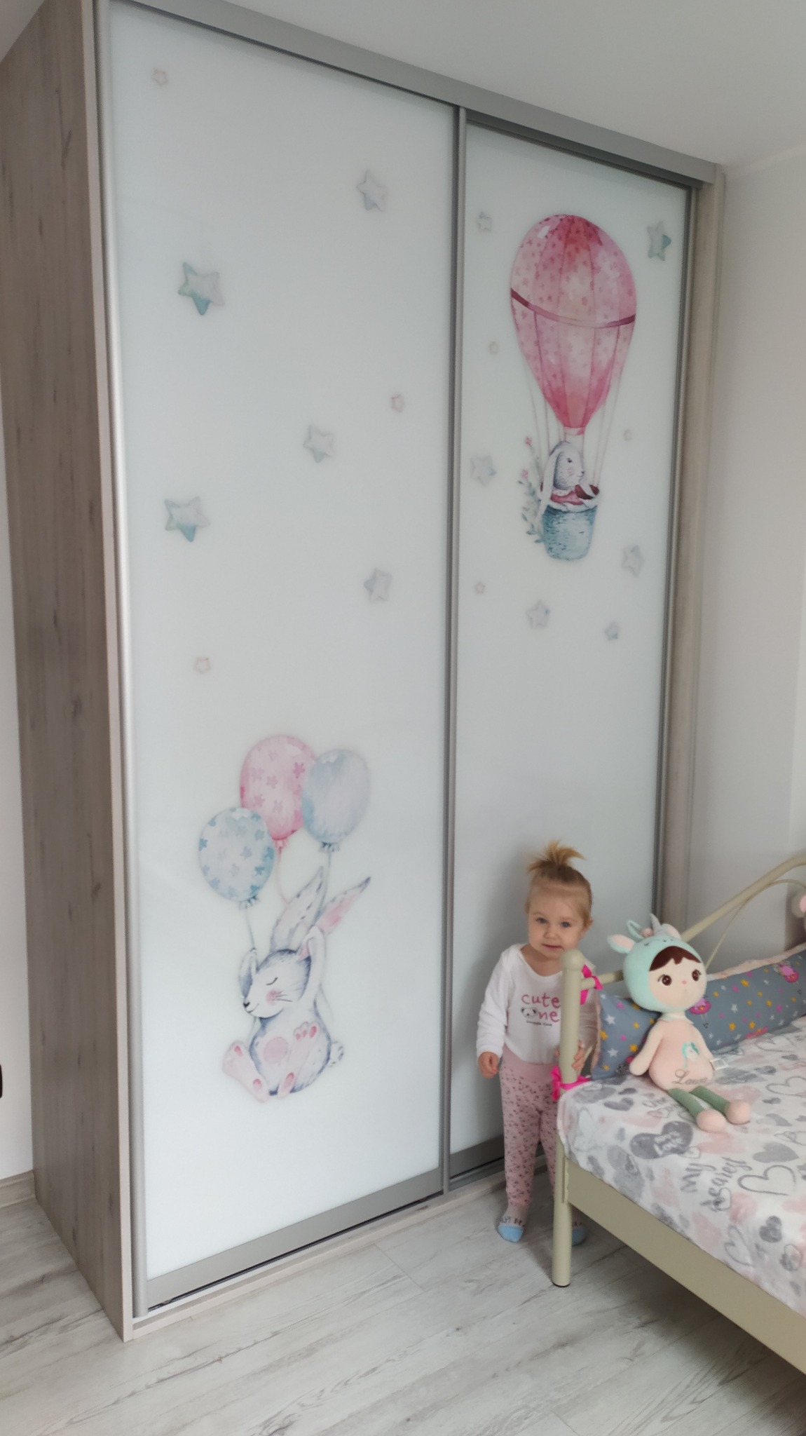 Dziecięca garderoba z przesuwnymi drzwiami, ozdobiona rysunkami królika z balonami i balonu na gorące powietrze, obok stoi mała dziewczynka.