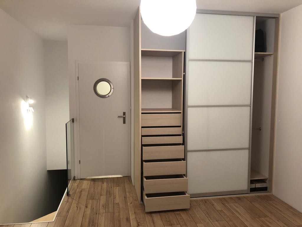 Nowoczesna zabudowa garderoby z przesuwnymi drzwiami i otwartymi półkami, widoczna na tle drewnianej podłogi i białych ścian z okrągłym oknem w drzwiach.