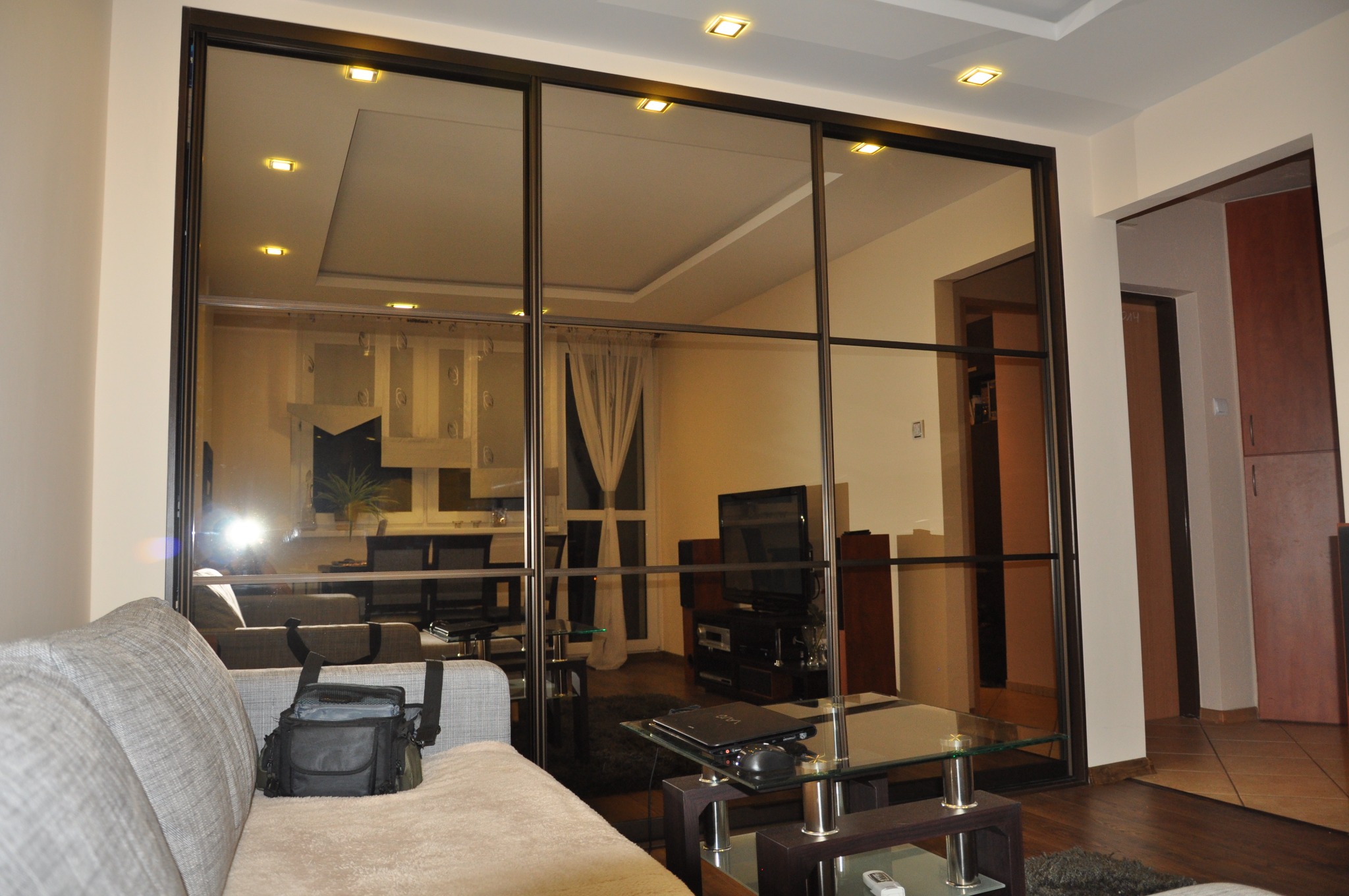 Duża, lustrzana szafa przesuwna z ciemnymi profilami, odbijająca wnętrze salonu z sofą, stolikiem kawowym i telewizorem.