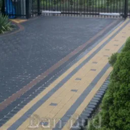 Ułożona nawierzchnia z kostki brukowej w kolorach szarości i beżu, tworząca wzór pasów i okręgów na podjeździe, z widoczną bramą w tle i roślinnością po bokach.