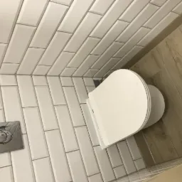 Nowoczesna toaleta wisząca z chromowanym przyciskiem spłukującym na ścianie z białych, prostokątnych płytek, uzupełnona podłogą imitującą drewno.