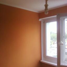 Remonty - Ściana pomalowana na odcień terakoty w pokoju z białym oknem i listwą przysufitową. Widoczna żarówka. Gładka powierzchnia, świeży efekt malowania.