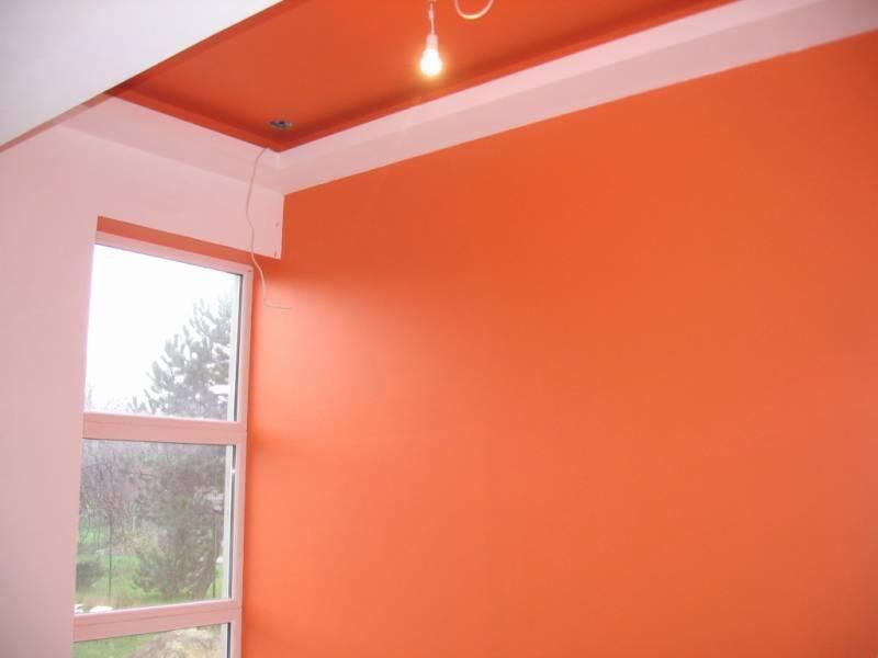 Pomarańczowa ściana w pokoju po malowaniu, widoczne okno z widokiem na zieleń, fragment sufitu z oświetleniem. Świeżo pomalowane powierzchnie.