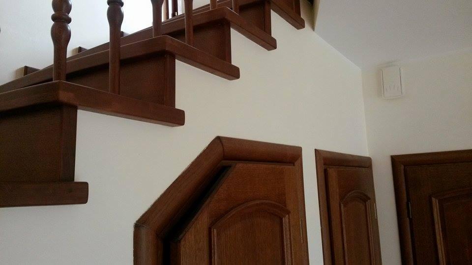 Klatka schodowa z drewnianymi schodami i balustradą, widok na pomalowaną na biało ścianę i dwoje drewnianych drzwi. Detal wykończenia wnętrza.