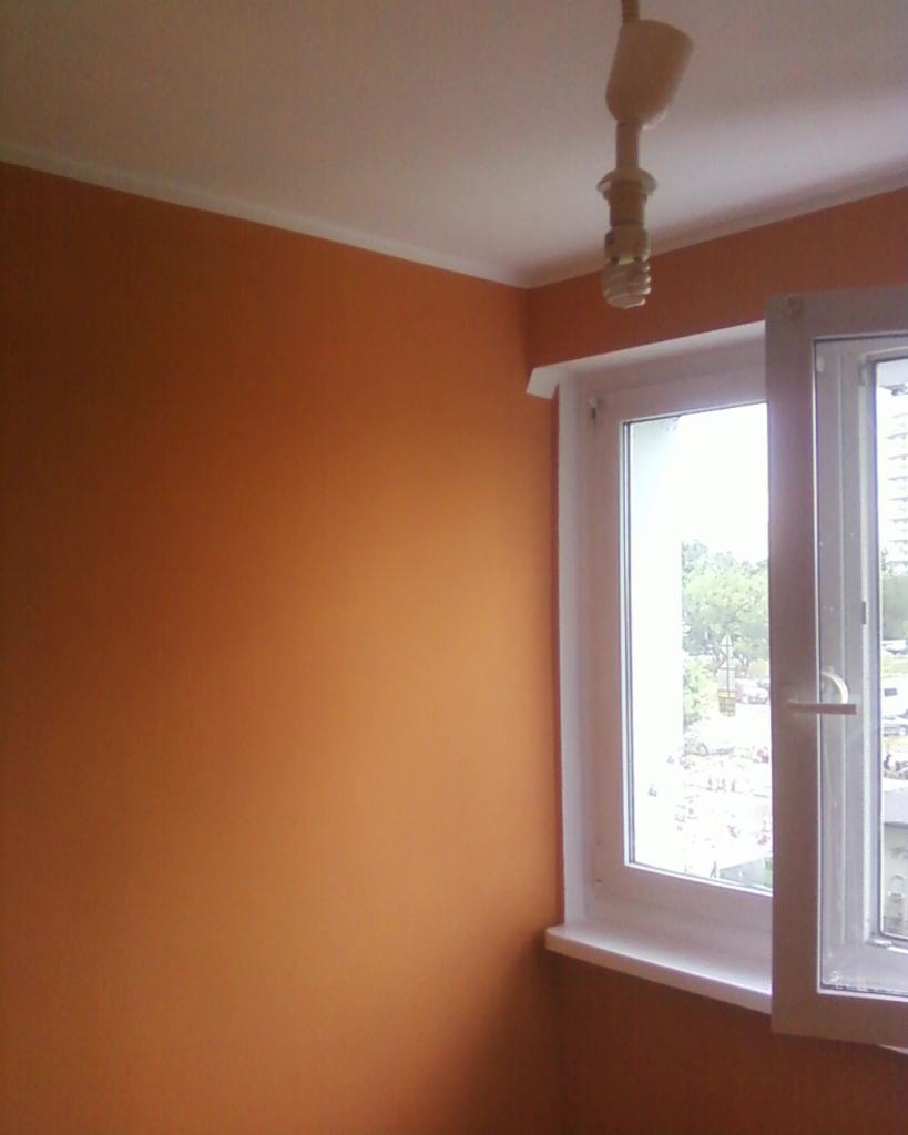 Ściana pomalowana na odcień terakoty w pokoju z białym oknem i listwą przysufitową. Widoczna żarówka. Gładka powierzchnia, świeży efekt malowania.