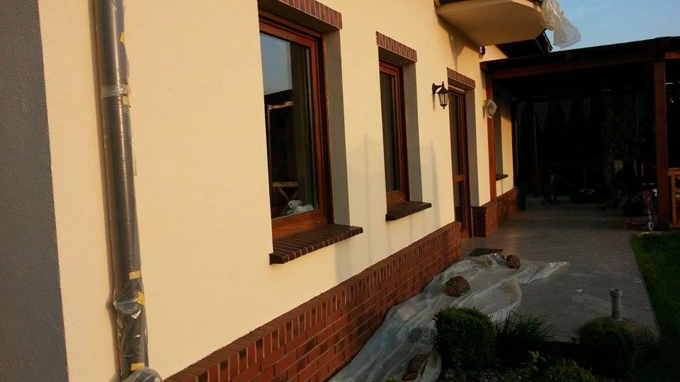 Elewacja domu po renowacji: kremowa ściana z brązowymi oknami i ceglaną podmurówką. Rura spustowa w folii ochronnej. Widok na taras.
