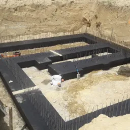 Przygotowany wykop pod fundamenty z izolacją pionową i wystającymi prętami zbrojeniowymi, widoczny pomarańczowy kubeł do betonu na łańcuchu, drabina oparta o skarpę wykopu.