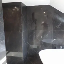 Wykończona łazienka z czarnymi, marmurkowymi płytkami na ścianach i podłodze, widoczny fragment białej wanny wolnostojącej z chromowaną armaturą.