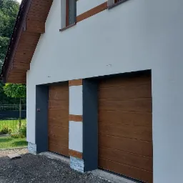 Nowoczesny dom z jasną elewacją, brązowymi akcentami przy oknach i bramie garażowej, oraz szarą kostką brukową przed budynkiem.