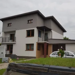 Dwukondygnacyjny dom z elewacją w odcieniach szarości i bieli, widoczny pracownik w trakcie malowania garażu oraz częściowo odsłonięty fundament z widocznymi uszkodzeniami.