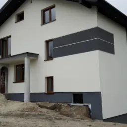 Dwukondygnacyjny dom z ocieploną elewacją w odcieniach bieli i szarości, widoczny z perspektywy narożnej, z przygotowanym terenem pod dalsze prace wykończeniowe.