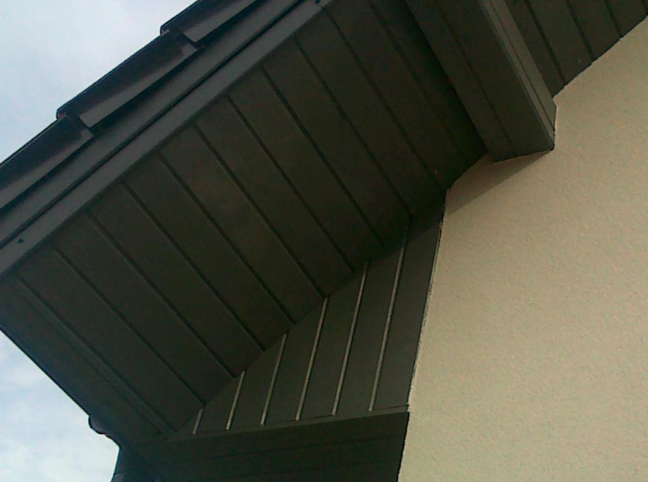 Ciemna, drewniana podbitka dachowa przy jasnej ścianie budynku, widok z dołu.