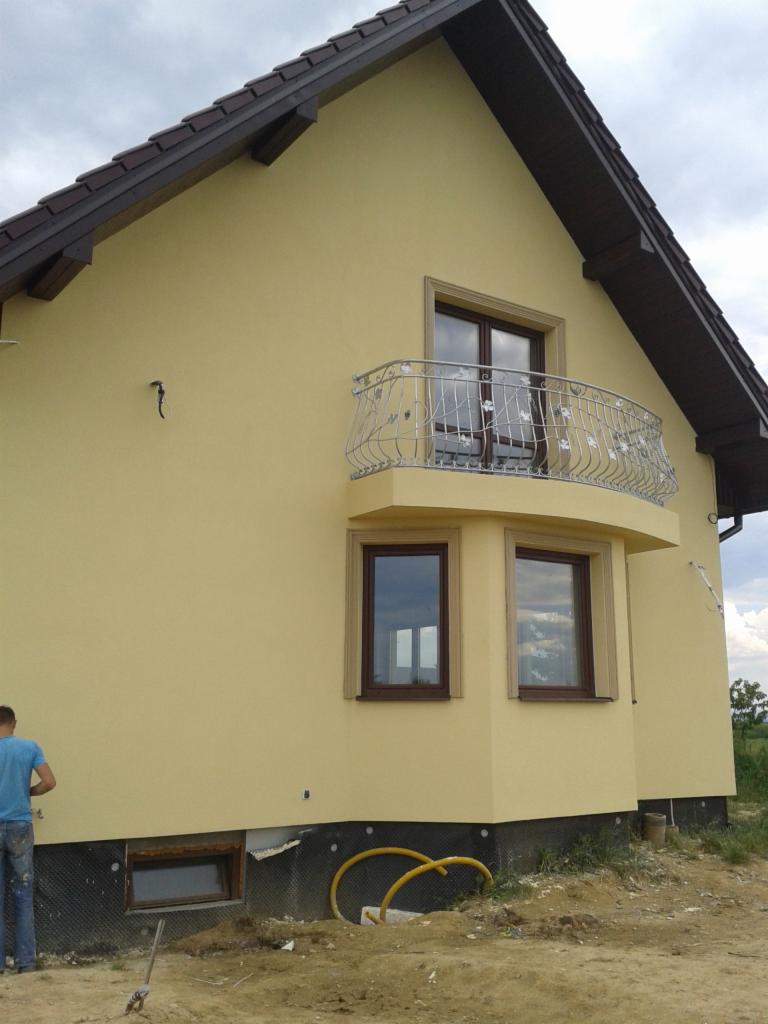Żółta elewacja domu z brązowymi oknami i balkonem z ozdobną balustradą. Widoczne fragmenty ocieplenia fundamentów i rury. Na pierwszym planie fragment sylwetki człowieka.