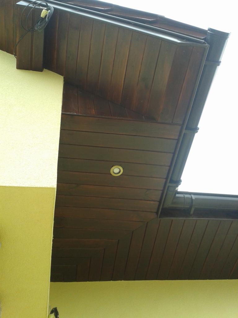 Ciemnobrązowa podbitka dachowa z wbudowanym punktowym oświetleniem, widok z dołu na fragment elewacji w kolorze jasnożółtym i system rynnowy.