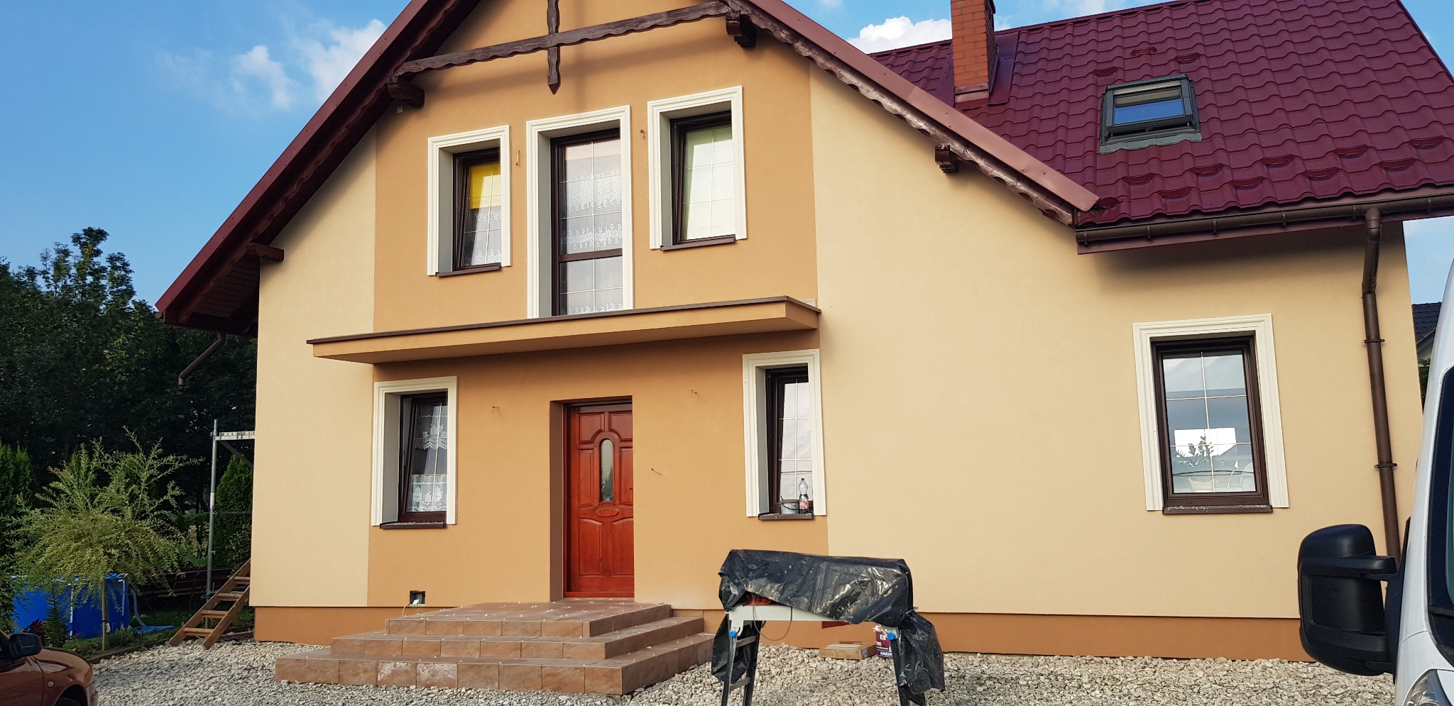 Dwukolorowa elewacja domu jednorodzinnego z czerwoną dachówką, widok z poziomu gruntu, z widocznymi schodami wejściowymi i przygotowanym stanowiskiem pracy z narzędziami pod folią.