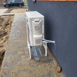 WACŁAWSKI DARIUSZ USŁUGI HYDRAULICZNE - Zewnętrzna jednostka klimatyzacji marki Hitachi zamontowana na metalowym stelażu przy ścianie budynku z szarą elewacją, widoczne kable w białej osłonie wchodzące do ściany, kostka brukowa...