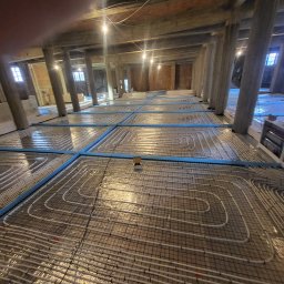 WACŁAWSKI DARIUSZ USŁUGI HYDRAULICZNE - Rozległa instalacja ogrzewania podłogowego w trakcie montażu w przestronnym, wysokim pomieszczeniu z drewnianą konstrukcją stropu i betonowymi kolumnami wsporczymi.