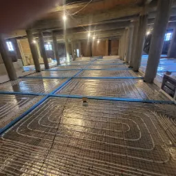 Rozległa instalacja ogrzewania podłogowego w trakcie montażu w przestronnym, wysokim pomieszczeniu z drewnianą konstrukcją stropu i betonowymi kolumnami wsporczymi.