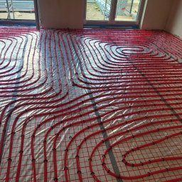 WACŁAWSKI DARIUSZ USŁUGI HYDRAULICZNE - Czerwone rury ułożone spiralnie na folii odblaskowej, tworzące system ogrzewania podłogowego w jasnym pomieszczeniu, widoczne okna i przygotowane elementy konstrukcyjne.