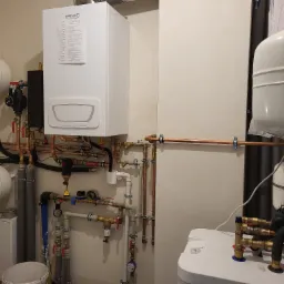 Kompletna instalacja grzewcza z białym kotłem gazowym, dwoma zbiornikami wyrównawczymi i rozbudowaną siecią rur miedzianych i plastikowych, widoczne liczne zawory i złączki.