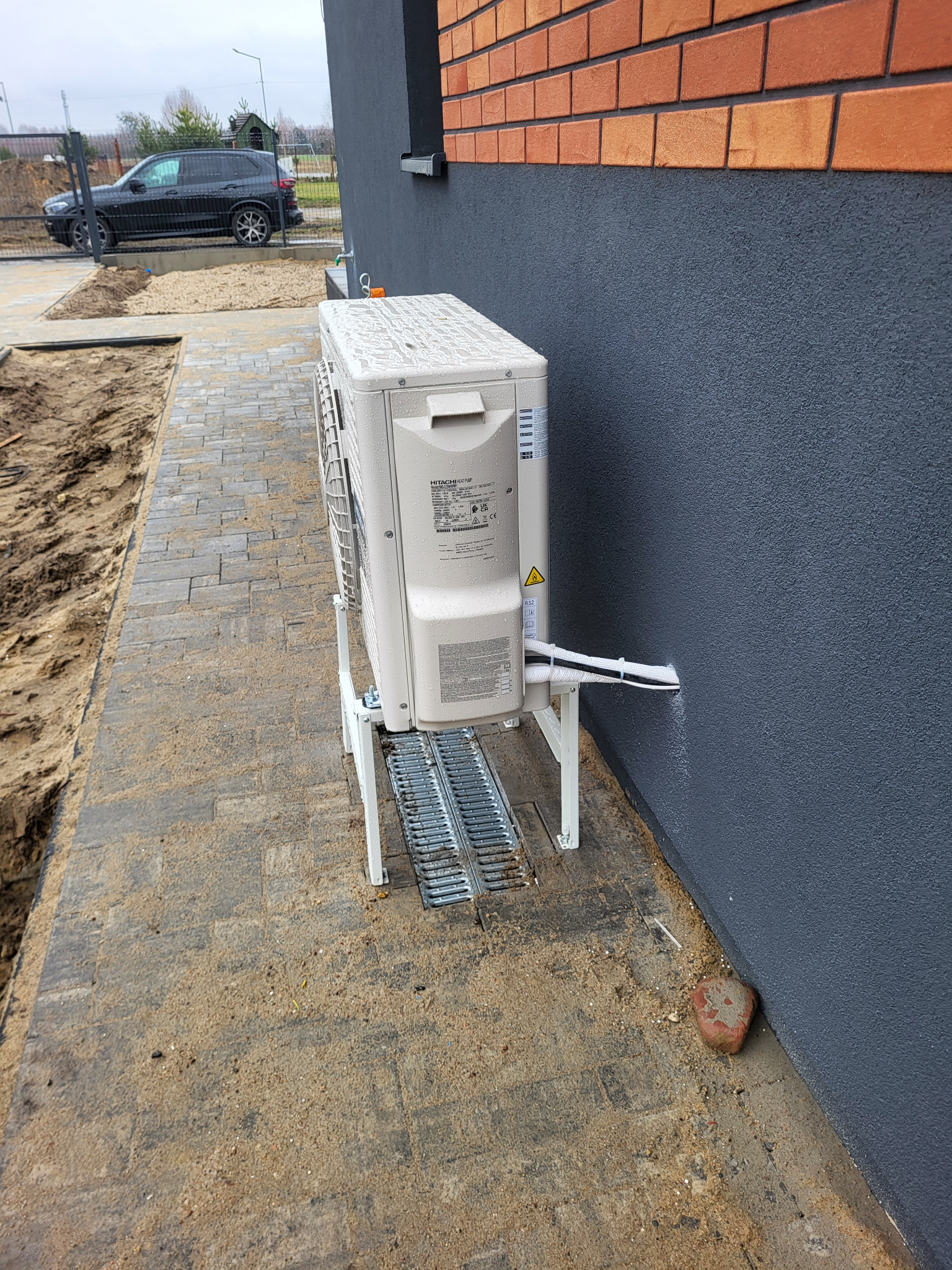 Zewnętrzna jednostka klimatyzacji marki Hitachi zamontowana na metalowym stelażu przy ścianie budynku z szarą elewacją, widoczne kable w białej osłonie wchodzące do ściany, kostka brukowa...