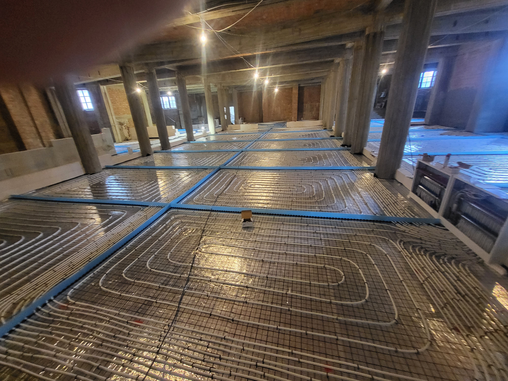 Rozległa instalacja ogrzewania podłogowego w trakcie montażu w przestronnym, wysokim pomieszczeniu z drewnianą konstrukcją stropu i betonowymi kolumnami wsporczymi.