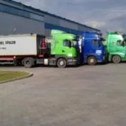 Rząd zaparkowanych ciężarówek w różnych kolorach (zielony, niebieski, czerwony) przed budynkiem magazynowym z błękitną elewacją. Na naczepie pierwszej ciężarówki widoczny napis 'TRANSPORT DANIEL...