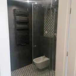 Nowoczesna łazienka z ciemnymi płytkami, grzejnikiem drabinkowym, kabiną prysznicową ze wzorzystymi płytkami wewnątrz i bidetem, widoczne przez otwarte drzwi.