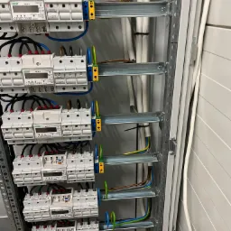 Szczegółowe ujęcie wnętrza rozdzielnicy elektrycznej z zamontowanymi licznikami i okablowaniem, widoczne białe liczniki F&F WZE-3 z wyświetlaczami, kolorowe przewody w peszlach i szyny montażowe.