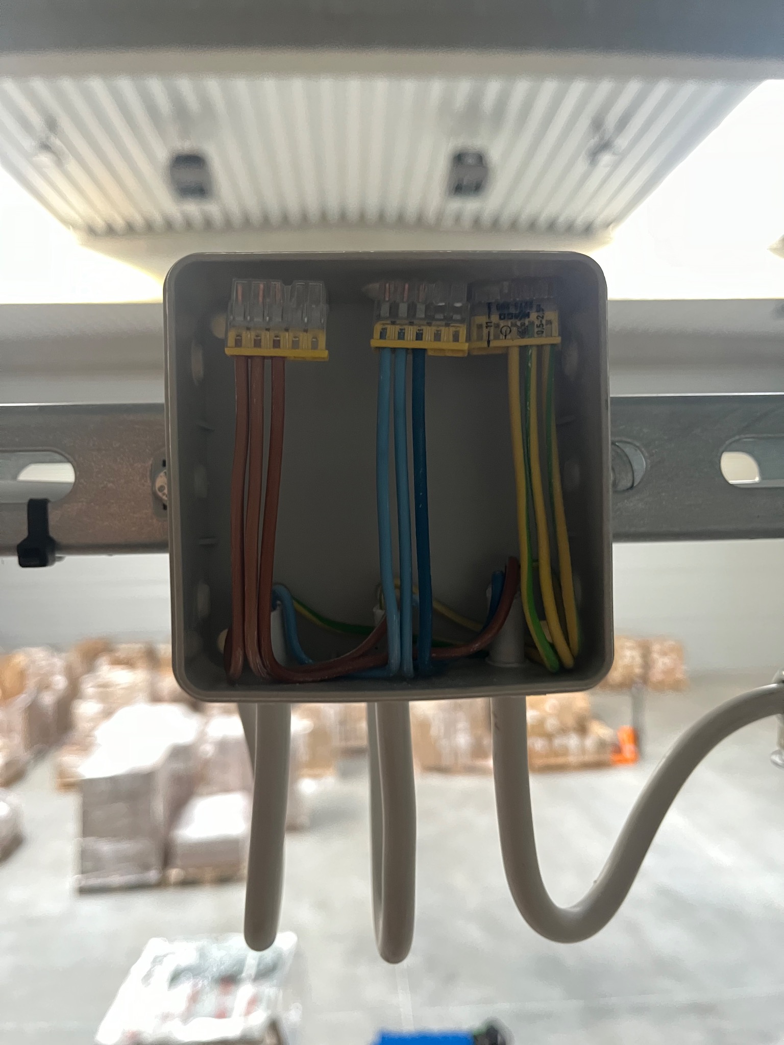 Szara puszka instalacyjna z podłączonymi przewodami (brązowe, niebieskie, żółto-zielone) za pomocą złączek WAGO, zamontowana na szynie w magazynie.