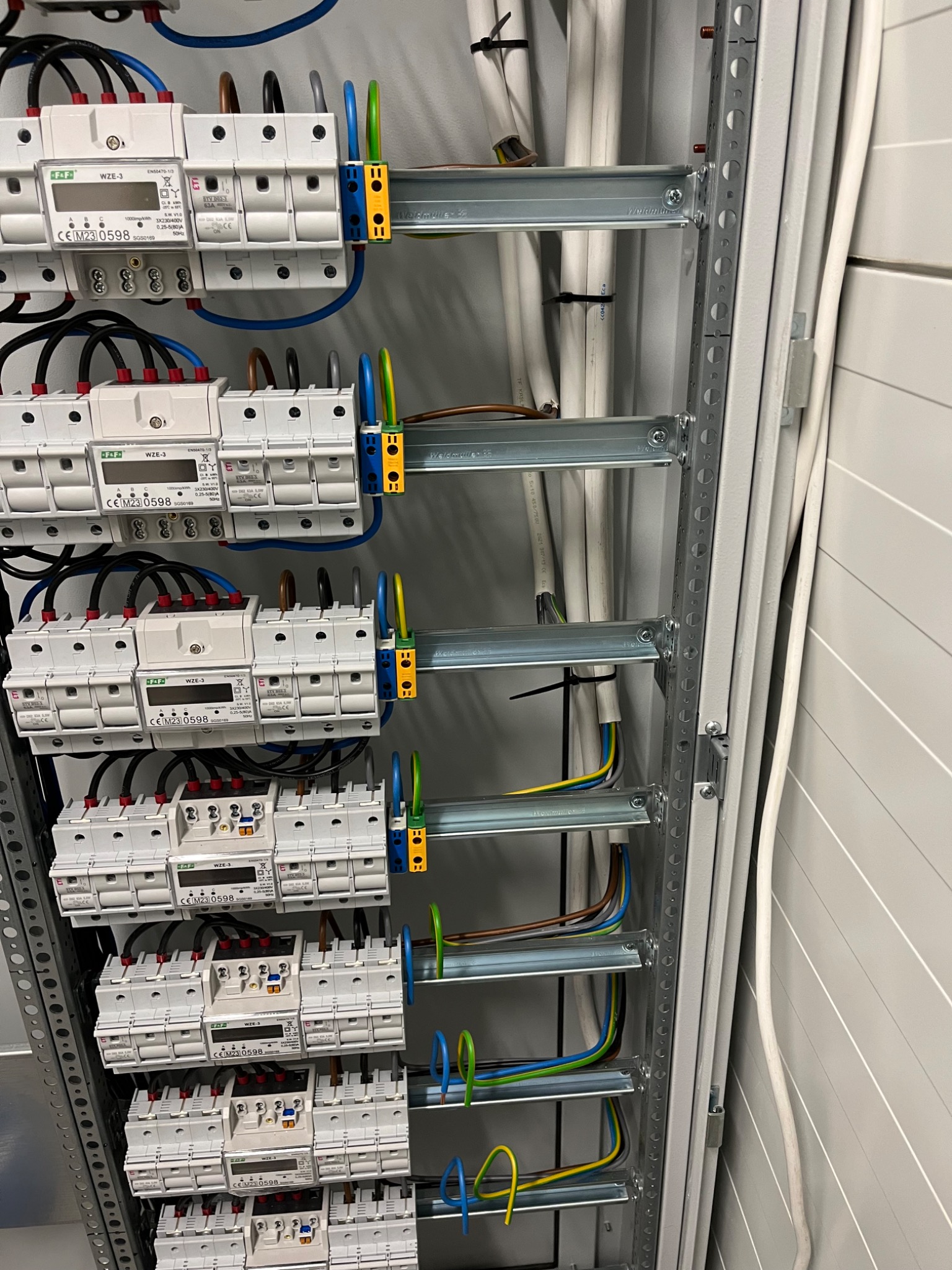 Szczegółowe ujęcie wnętrza rozdzielnicy elektrycznej z zamontowanymi licznikami i okablowaniem, widoczne białe liczniki F&F WZE-3 z wyświetlaczami, kolorowe przewody w peszlach i szyny montażowe.
