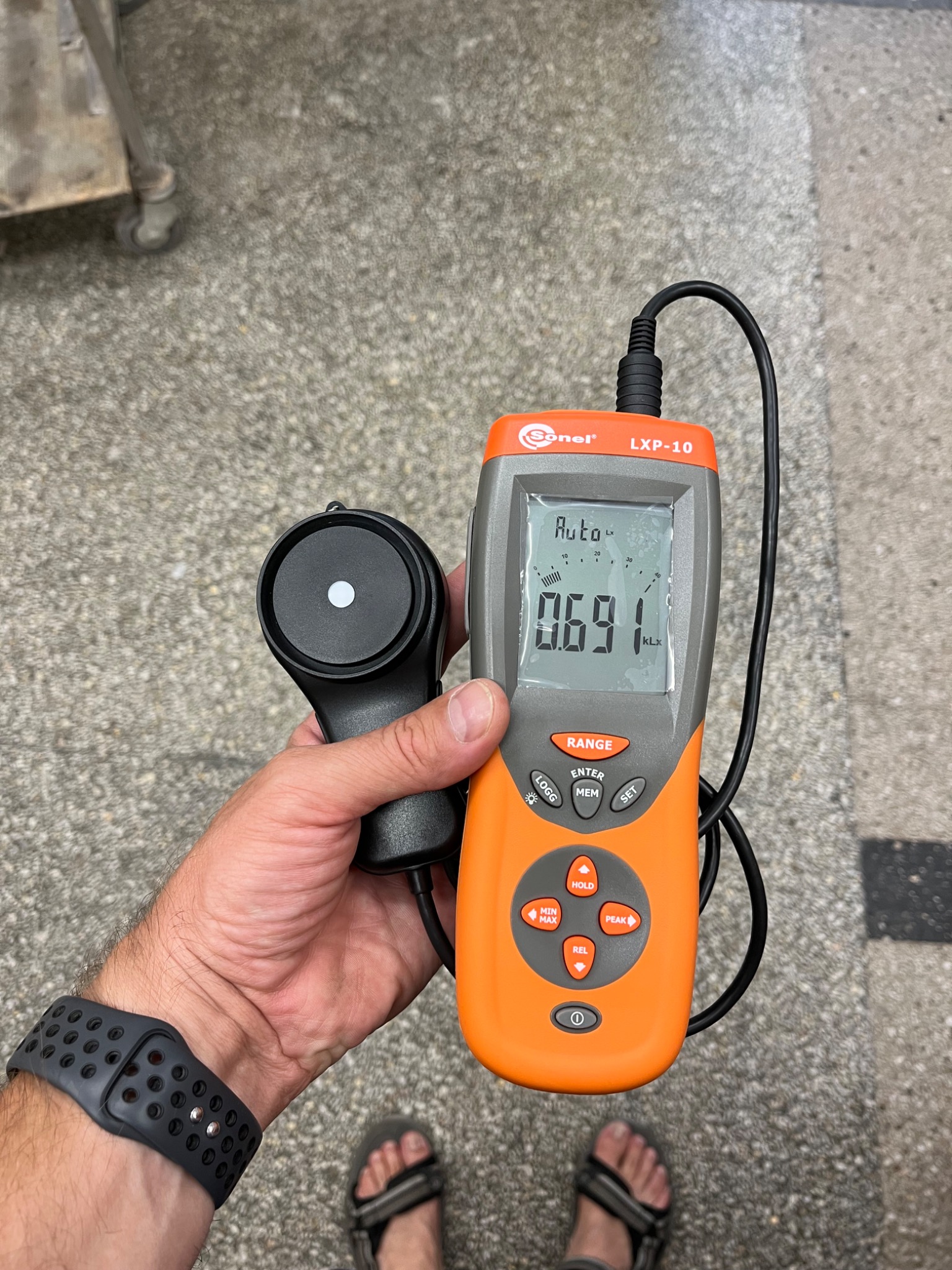 Miernik natężenia oświetlenia Sonel LXP-10 w dłoni, wyświetlający wartość 0.691 kLx, na tle betonowej posadzki i fragmentu wózka transportowego.