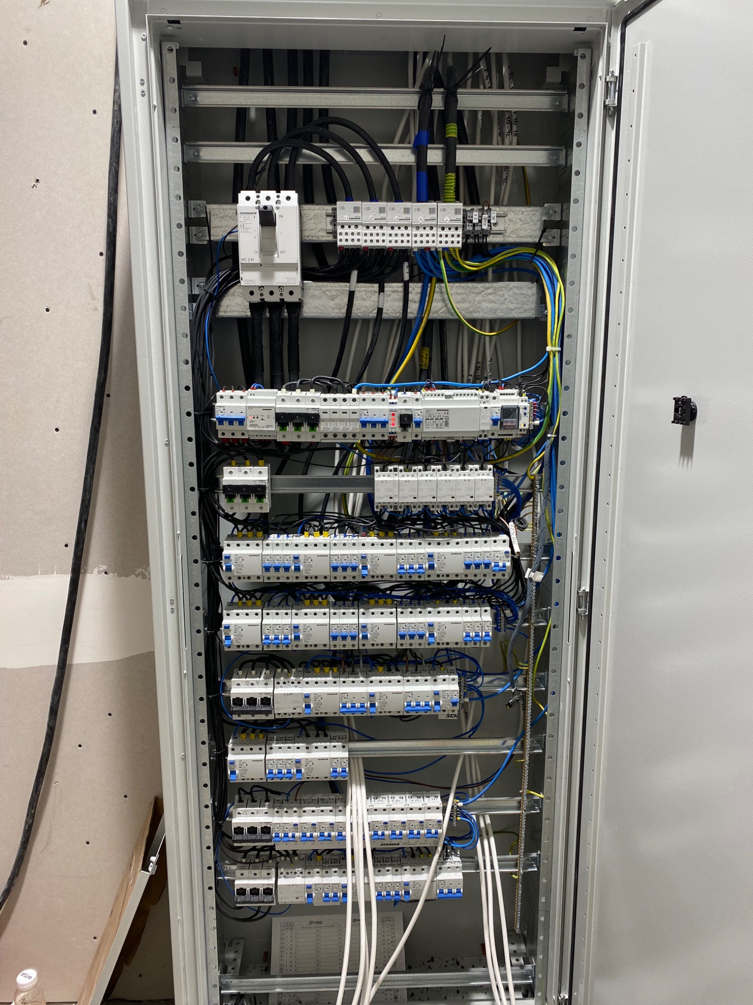 Otwarta szafa elektryczna z zamontowanymi bezpiecznikami, kablami i listwami, widoczne przewody w kolorach czarnym, niebieskim, żółto-zielonym i białym, ściana w tle przygotowana do malowania.