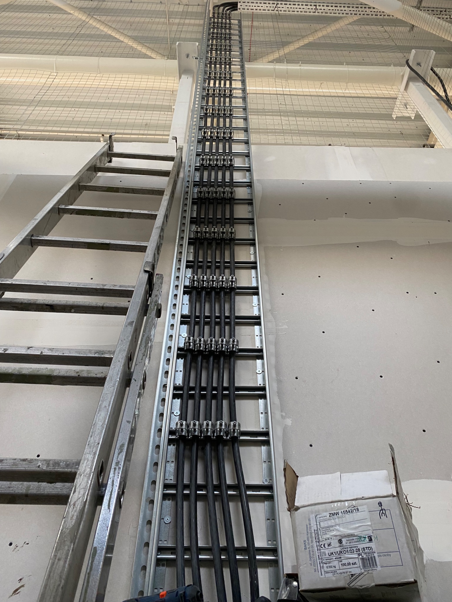 Montaż przewodów elektrycznych w metalowej korycie kablowej na ścianie w surowym stanie, widoczna drabina aluminiowa i karton z uchwytami kablowymi.