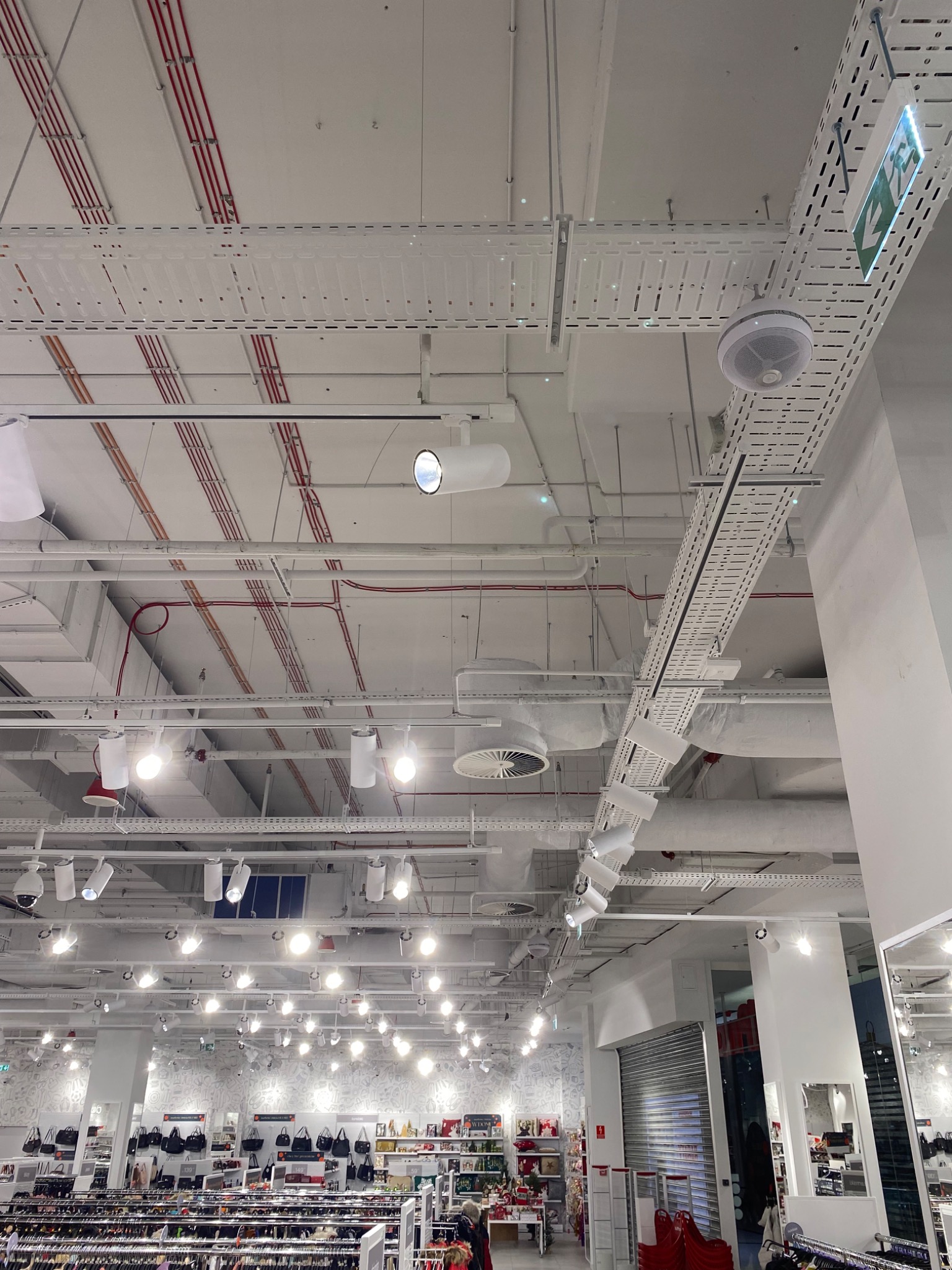 Sufit w sklepie z widocznymi instalacjami: korytka kablowe, rury w kolorze czerwonym, oświetlenie punktowe i głośniki sufitowe, oraz znak ewakuacyjny.
