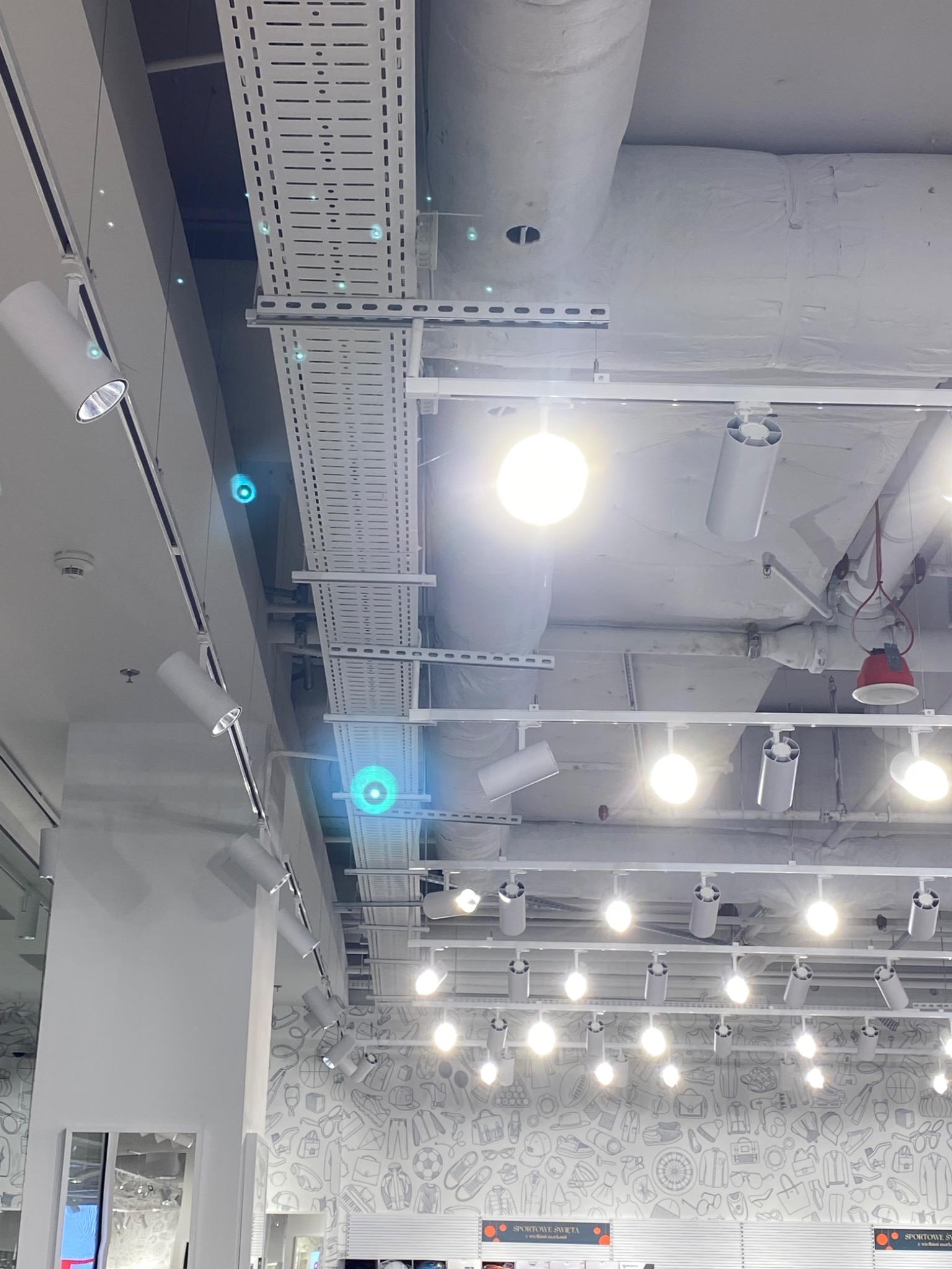 Białe, nowoczesne oświetlenie punktowe na szynoprzewodach pod sufitem w sklepie, widoczne koryta kablowe i instalacje sufitowe.