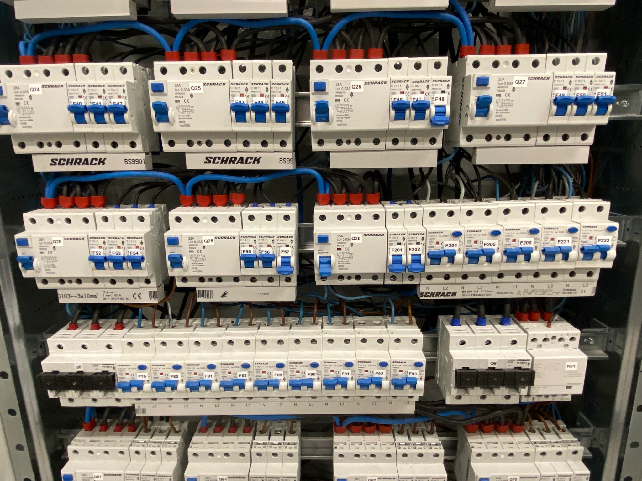 Rozdzielnia elektryczna z licznymi bezpiecznikami Schrack, okablowanie w kolorach niebieskim i czerwonym, widoczne oznaczenia faz i numeracja poszczególnych elementów.