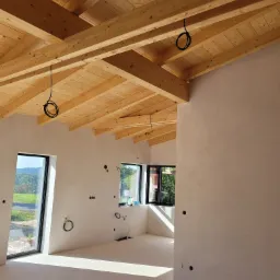 Jasne wnętrze w trakcie wykańczania: świeżo otynkowane ściany, drewniany strop z widocznymi kablami elektrycznymi, okna z widokiem na zieleń za oknem.