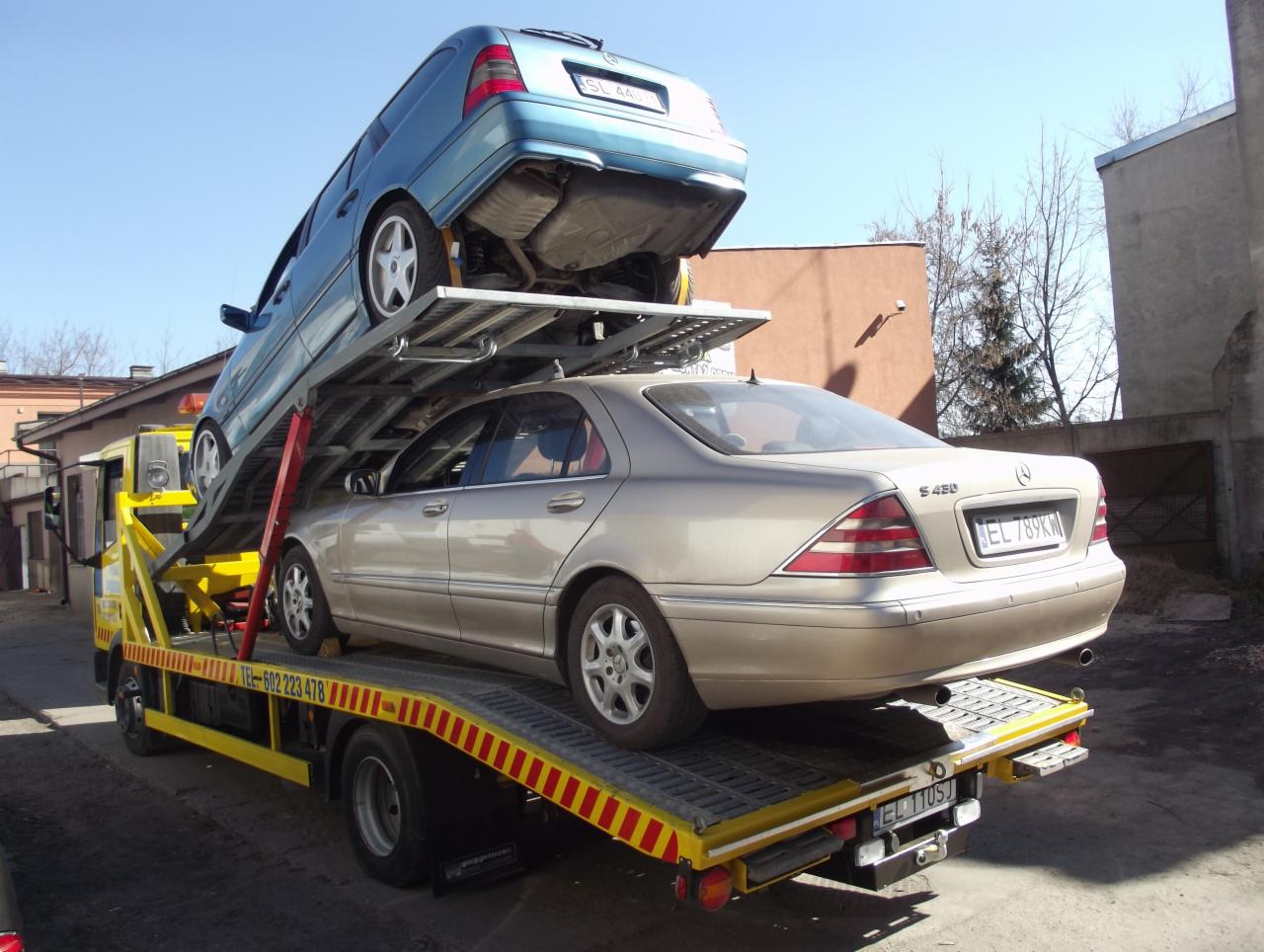 Żółta laweta z dwoma samochodami osobowymi: niebieskim na górze i beżowym na dole, widoczne numery rejestracyjne. Słoneczny dzień.