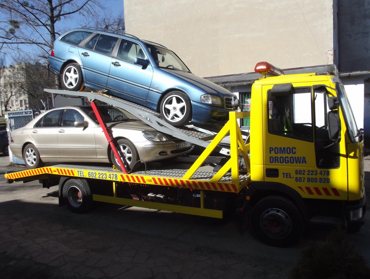 Żółta laweta z dwoma samochodami (niebieskie kombi i beżowa limuzyna) na platformie. Widoczne numery telefonów na kabinie. Słoneczny dzień, budynek w tle.
