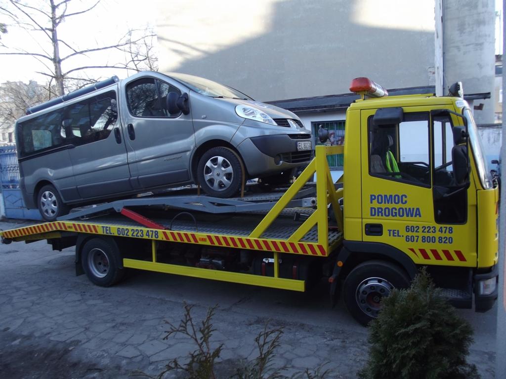 Żółta laweta z szarym busem na platformie. Widoczne numery telefonów. Pojazd na tle miejskiej zabudowy.