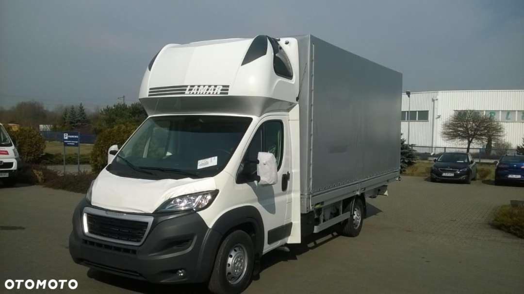 Biały bus dostawczy z zabudową plandekową i napisem LAMAR na dachu kabiny, zaparkowany na placu przed budynkiem w słoneczny dzień. Widok z boku.