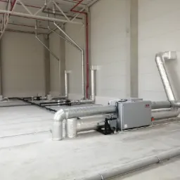 Szara centrala wentylacyjna SALDA z izolowanymi przewodami w hali przemysłowej, widoczne rury pod stropem i instalacja przeciwpożarowa.