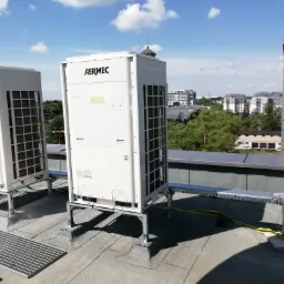 Dwa białe klimatyzatory Aermec MVA na dachu budynku z widokiem na miasto w tle, zamontowane na metalowych konstrukcjach.