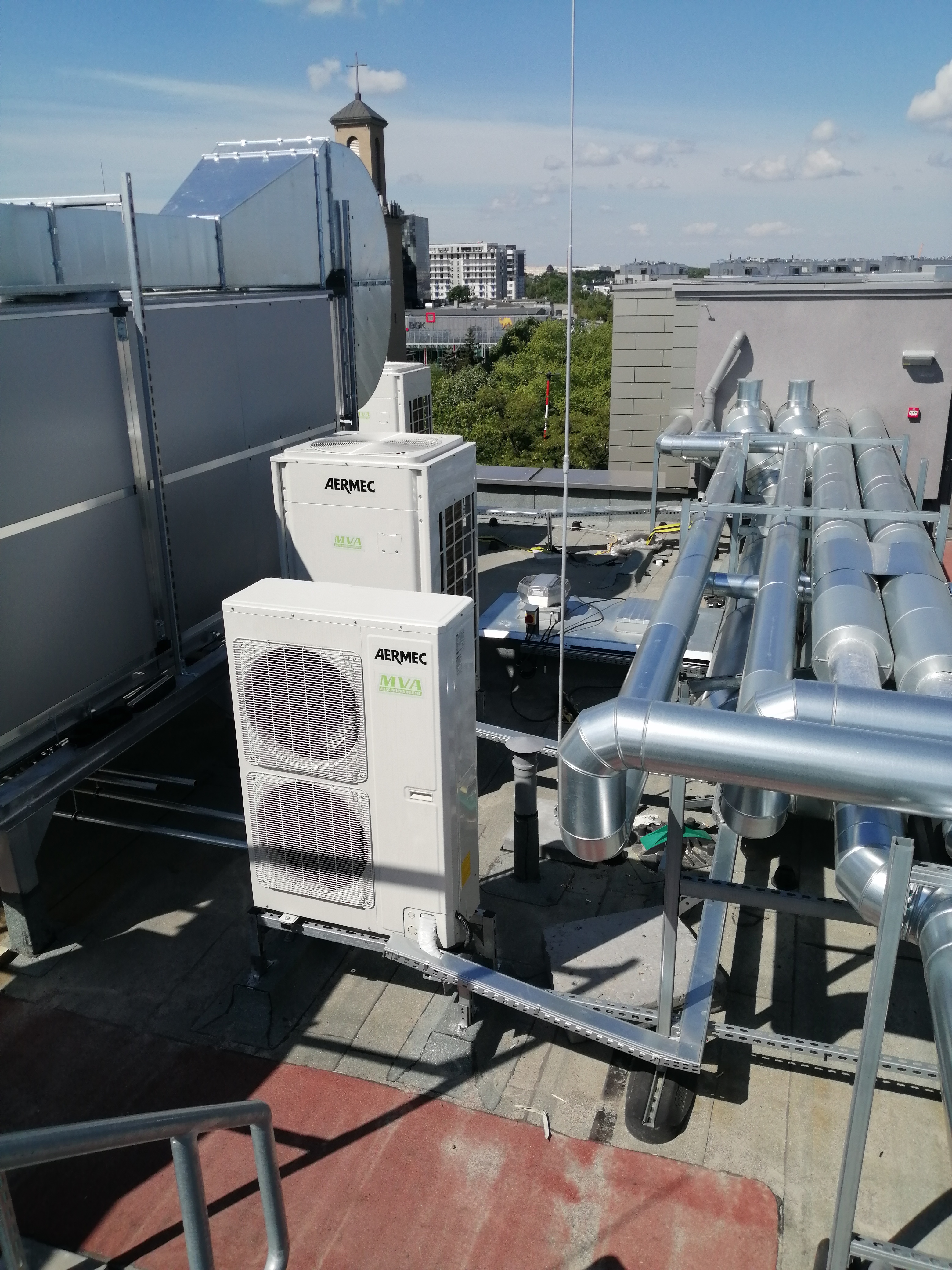Instalacja klimatyzacji przemysłowej Aermec MVA na dachu budynku z widocznymi rurami wentylacyjnymi i fragmentem panoramy miasta w tle.