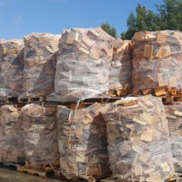 Rząd ułożonych palet z workami pociętego drewna opałowego, zabezpieczonych folią, na tle zielonych drzew i błękitnego nieba.