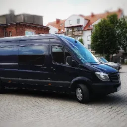 Czarny bus Mercedes-Benz Sprinter na parkingu z kostki, budynki w tle. Pojazd o podwyższonym dachu, z długim rozstawem osi, idealny do transportu osób lub towarów.